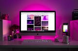 An iMac desktop set-up with warm, soft pink lighting coming from the back of the monitor. The desk has a coffee mug, smartphone and mouse to the right, wireless keyboard in the front, and a little bonsai and other small table decor to the left. It looks clean and well organised.