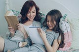 A mother and daughter relax together on a bed, reading, with smiles on their faces.