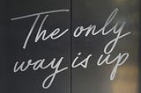 Steel elevator doors prepare to open so you can rise to greater elevation. Written in upbeat cursive across the metal: “The only way is up.”