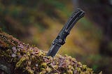 A black Ruger compact knife stuck onto a horizontal moss covered tree trunk. The handle looks ergonomic and comfortable to hold.