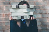 A woman holding books up to obscure her face. She stands in front of a brick wall.