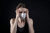 Woman with a face mask on, holding her temples. She has a bad cold.