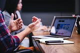 A person is speaking to others around the table as their laptop is open to a project page an their phone sits on top of their notepad in front of it.