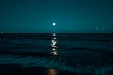 Photo of a beach at night, under a full moon. The sky is shades of dark turquiose, and the water looks darker with the night. A gentle wave meets the sandy beach with white motion in the dark night.