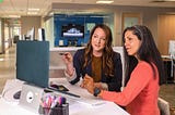 Two women are talking by a desk. They are looking at a computer screen.