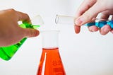 Someone is pouring a beaker of green liquid, and one of blue liquid, into a flask of red liquid.