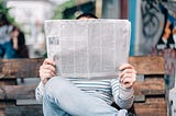 A photo of someone sitting on a wooden park bench, with their legs crossed, holding up a broadsheet newspaper. The newspaper covers their face; only the top of their head, with short black hair, is visible