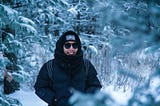 Smiling person in dark jacket, hood, and glasses, in front of snow-covered trees