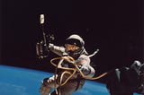A person in a space suit holding a large tool is floating above the earth, tethered to a space craft with by a twisted cord.