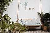 A swing bench with white pillows on it, surrounded by plants. The words “Self Love” are written on the white wall behind it.