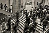 This image depicts a bustling interior scene, possibly within a museum or public building, characterized by a sense of motion and activity. The central focus is a grand staircase populated by numerous individuals depicted in various states of motion blur, which suggests the dynamic flow of people moving up and down. Some individuals are captured in stillness, offering contrast to the blurred motion around them, which gives the scene a temporal dimension. The architecture features classic stone w