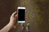 A person standing on a trail in the woods (view only of legs with feet clad in socks and sneakers ) looking at an empty screen on a smartphone.