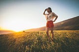 Young woman standing in a field on a sunny day shielding her eyes with her hand while looking out into the distance across the field.