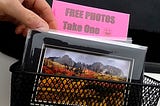 A hand reaching into a bin filled with 5x7 matted photos with a sign that says “Free Photos — Take One”