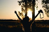 A woman faces the sun, holds a camera in her right hand, and spreads her arms widely