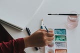 Person sitting at a desk with a sketchbook painting with water colours.