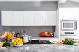 a kitchen with whute cabinets and a grated stove and a marble counter. there is a bowl of lemons on the counter and orange pots and pans