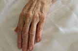 The wrinkled, blue veined hand of an old woman lies atop white sheets.