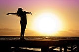 Silhouette of woman standing on a dock facing the sun with her arms outstretched at golden hour