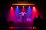 A white man with a beard wearing a white collared shirt, red scarf, and dark pants stands on a stage backlit by alternating blue and red lights in an otherwise dark room. He has his arms slightly outstretched. The dark silhouettes of a few people in the audience can be seen.