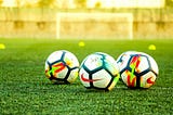 Three soccer balls sit on a turf soccer field with small yellow cones and a goal blurred in the background.