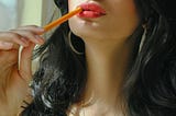Close-up of a brunette wearing red lipstick chewing on a pencil.