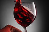 A full glass of red wine, held at the stem by a person’s fingertips, is sloshed over the edge.
