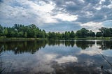 A lake surrounded by trees reflects a blue sky and clouds.