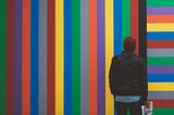 A man holding a newspaper stares at a wall painted with brightly colored vertical stripes