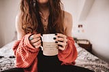A person sits on a bed while holding a coffee cup that reads “dear life, it’s beautiful here.”