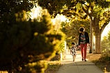 A mother walks her 2 year old daughter down a sidewalk in a sunny afternoon shaded by trees.