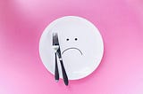 An empty plate with a sad face drawn on it. With a fork and knife on top of the plate.