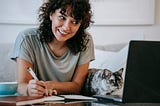 Smiling woman at home with cat writing in planner while using laptop