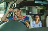 A mom, looking exhausted is sitting in her car, looking down, weighed down with trouble, her daughter sits in the back seat looking on