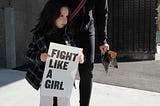 A little girl holding a placard that says “Fight Like A Girl”. The mother is standing beside and holding the girl.