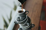 A stack of ceramic coffee mugs stands precariously as someone pours hot coffee from a carafe into the top mug.