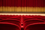 A theater. Perspective from the audience. Rows of empty red velvet chairs and in the distance, a red curtain with gold trim.