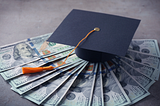 Navy blue graduation cap with orange tassle on a stack of $100 bills