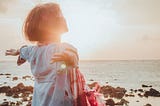 Lady standing on the shore with outstretched arms, and sunlight shining on her.