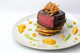 A medium rare steak fillet cut to show the inside. It is beautifully plated with dabs of orange and yellow sauce around a white plate. Delicious-looking fried onions are piled atop the meat. Yum!