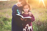 Two little girls sharing the joy of a book while standing in a sunlit field.