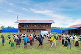 A group stretches arms towards the left in rows, a morning exercise in a rehabilitation program. They stand in the center of a field with the rehabilitation center behind them.