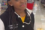 Nya Jingles is a 15 year old teen. She is an African American girl. She is seen in a grocery store with her hair in braids, a yellow shirt on and a black and white letterman jacket.