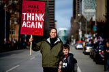 A man and child holding up a sign that says “Make America Think Again”