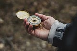 A person’s hand holds an old, gold compass, against a backdrop of autumn leaves and layered sleeves.