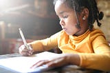 Black girl writing with a pen on paper. She is wearing a yellow shirt and has braids in her hair. She appears to be concentrating.
