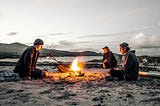 3 men sitting around a survival camp fire.