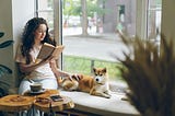 a woman reads a book next to a window with a coffee and her dog by her side
