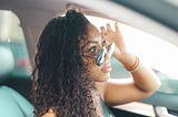 Lady with long curly hair looking over sunglasses while driving.