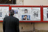 A man in a tweed jacket reads the daily newspaper. It’s protected behind glass, set up along an outdoor wall for all to read.